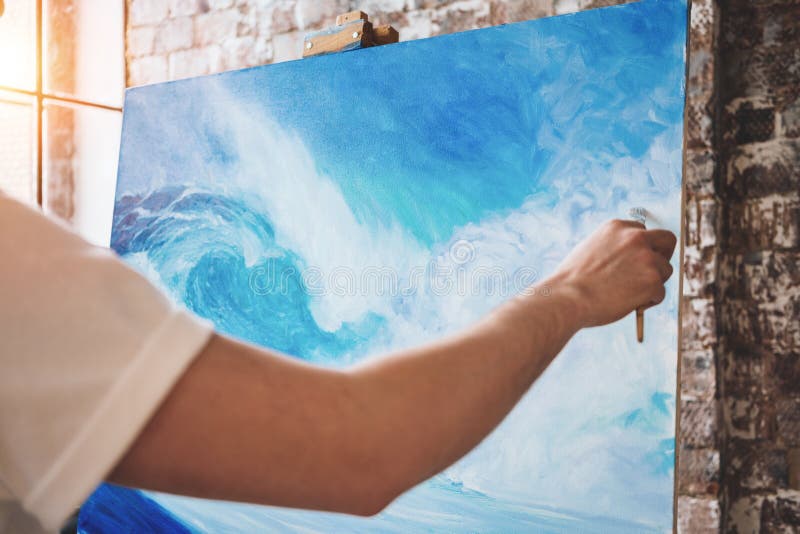 Male hand of artist closeup holding paintbrush on background of canvas on easel. Professional painter drawing ocean wave in loft studio. Flare effect. Male hand of artist closeup holding paintbrush on background of canvas on easel. Professional painter drawing ocean wave in loft studio. Flare effect