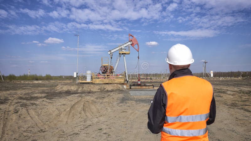 Męski operator w sygnałowej kamizelce i białym hełmie nagrywa czytania o pompuje jednostka oleju, benzynowy i opałowy obraz stock Męski operator w sygnałowej kamizelce i białym hełmie nagrywa czytania o pompuje jednostka oleju, benzynowy i opałowy obraz stock
