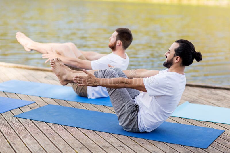 Fitness, sport, yoga, people and healthy lifestyle concept - men making half-boat pose on mat outdoors on river or lake berth. Fitness, sport, yoga, people and healthy lifestyle concept - men making half-boat pose on mat outdoors on river or lake berth