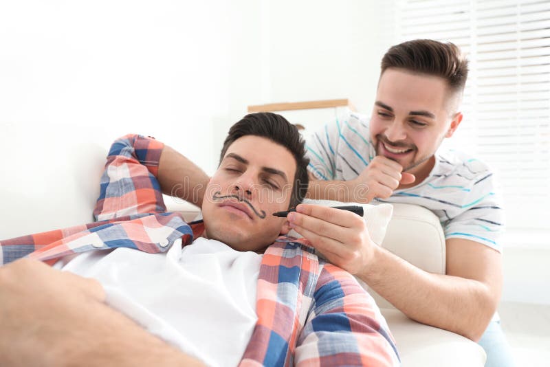 Mężczyzna rysujący wąsy na twarzy śpiącego przyjaciela w domu. dzień kwiecieli