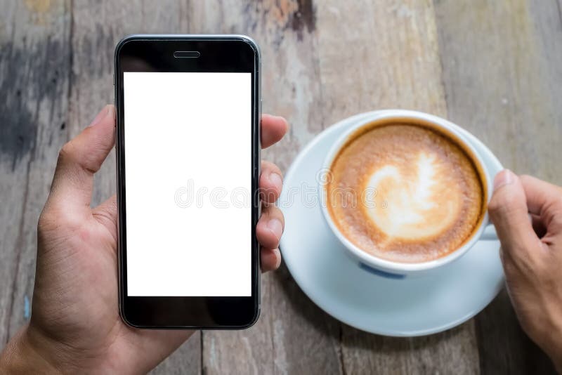 Mężczyzna ręka trzyma mądrze telefon gorący z latte, filiżanka na drewnianym biurku w sklepie z kawą Smartphone z pustym ekranem obraz royalty free Mężczyzna ręka trzyma mądrze telefon gorący z latte, filiżanka na drewnianym biurku w sklepie z kawą Smartphone z pustym ekranem obraz royalty free