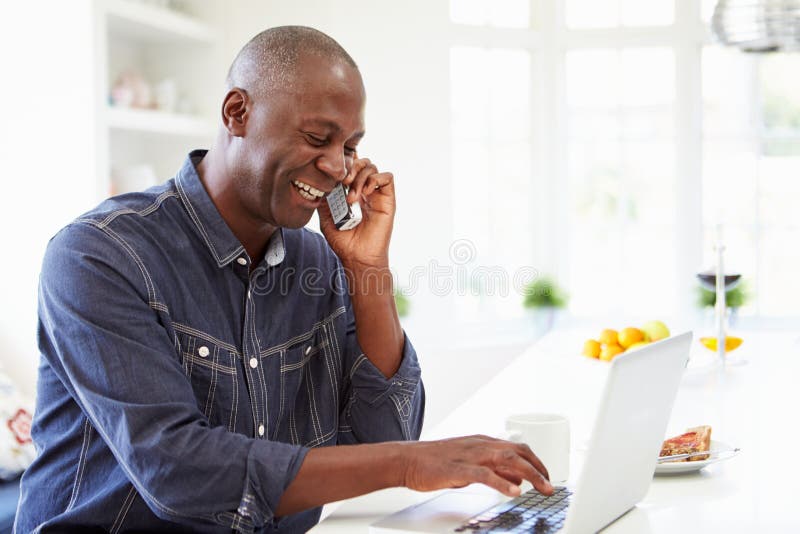 Mężczyzna Używa laptop I Opowiadający Na telefonie W kuchni W Domu zdjęcia stock Mężczyzna Używa laptop I Opowiadający Na telefonie W kuchni W Domu zdjęcia stock