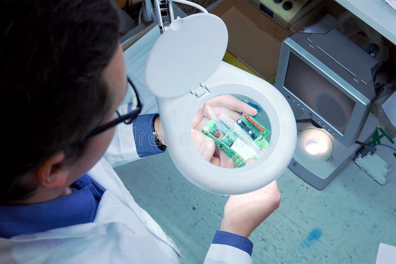 Mężczyzna w białym kontuszu i szkłach sprawdza elektryczną deskę dla defektów przez powiększać - szkło Kontrola jako?ci zdjęcia stock Mężczyzna w białym kontuszu i szkłach sprawdza elektryczną deskę dla defektów przez powiększać - szkło Kontrola jako?ci zdjęcia stock