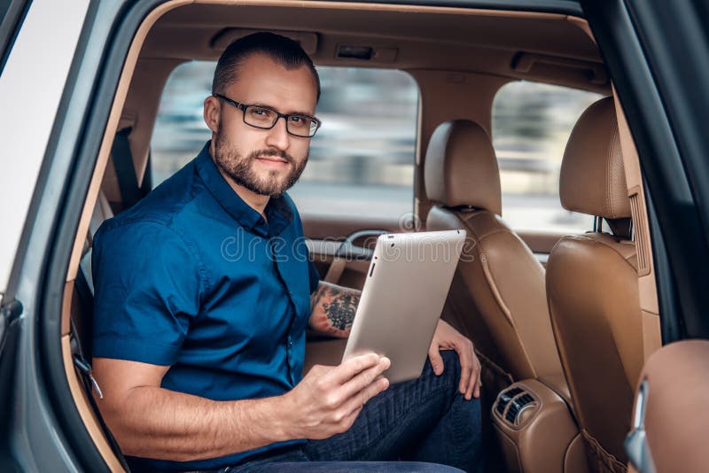 Mężczyzna w eyeglasses z tatuażem na jego ręce używać przenośnego pastylka peceta na tylnym siedzeniu samochód zdjęcia royalty free Mężczyzna w eyeglasses z tatuażem na jego ręce używać przenośnego pastylka peceta na tylnym siedzeniu samochód zdjęcia royalty free