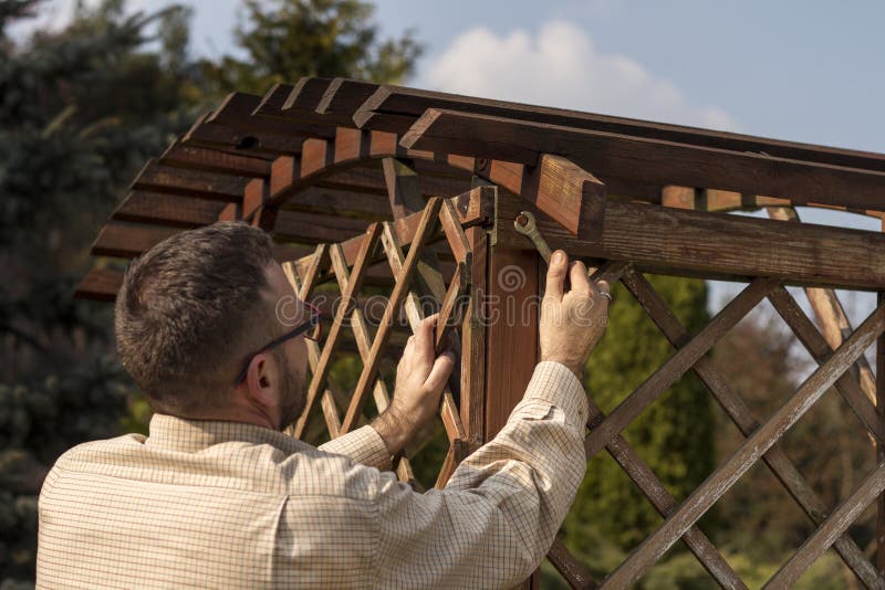 A man with glasses and a flannel shirt during spring gardening. Renovation and maintenance of garden pergolas. The man`s hand tightens the screw with a wrench. A man with glasses and a flannel shirt during spring gardening. Renovation and maintenance of garden pergolas. The man`s hand tightens the screw with a wrench