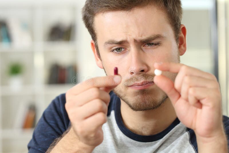 Portrait of a man holding medicines wondering about a round white pill or red capsule sitting on a couch in a house interior. Portrait of a man holding medicines wondering about a round white pill or red capsule sitting on a couch in a house interior