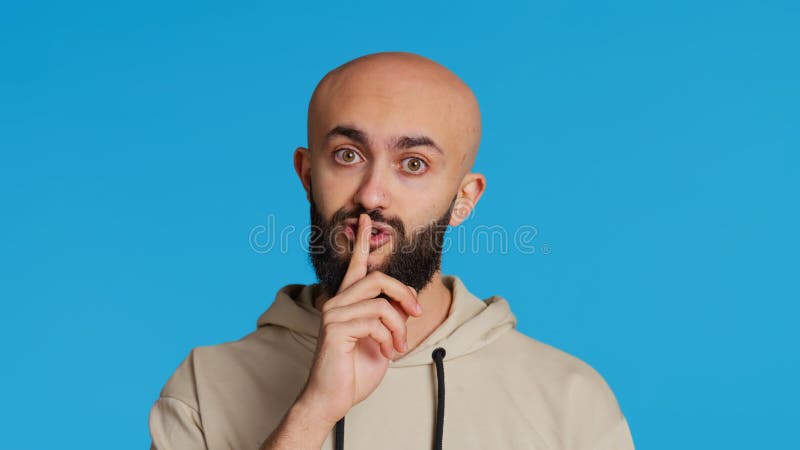 Middle eastern man doing hush silence symbol in studio, asking to keep a secret over blue background. Arab person creating mute gesture to be quiet in front of camera. Camera 2. Middle eastern man doing hush silence symbol in studio, asking to keep a secret over blue background. Arab person creating mute gesture to be quiet in front of camera. Camera 2.