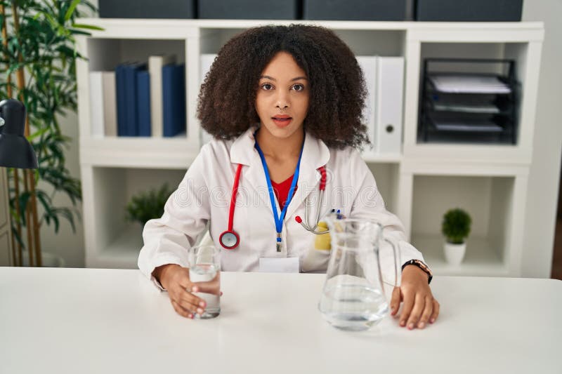 Young african american doctor woman drinking water at the clinic scared and amazed with open mouth for surprise, disbelief face. Young african american doctor woman drinking water at the clinic scared and amazed with open mouth for surprise, disbelief face