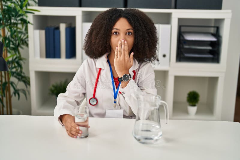 Young african american doctor woman drinking water at the clinic covering mouth with hand, shocked and afraid for mistake. surprised expression. Young african american doctor woman drinking water at the clinic covering mouth with hand, shocked and afraid for mistake. surprised expression