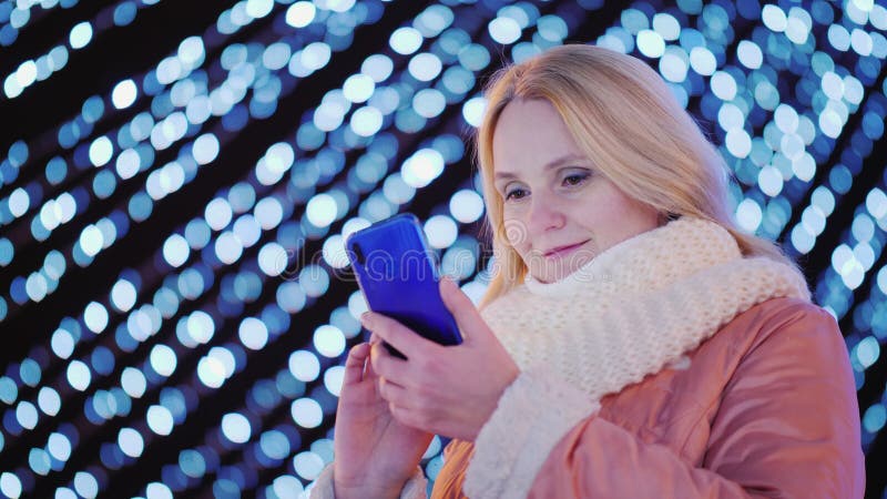 Młoda atrakcyjna kobieta używa smartphone na tle świąteczne girlandy w mieście zdjęcie stock Młoda atrakcyjna kobieta używa smartphone na tle świąteczne girlandy w mieście zdjęcie stock