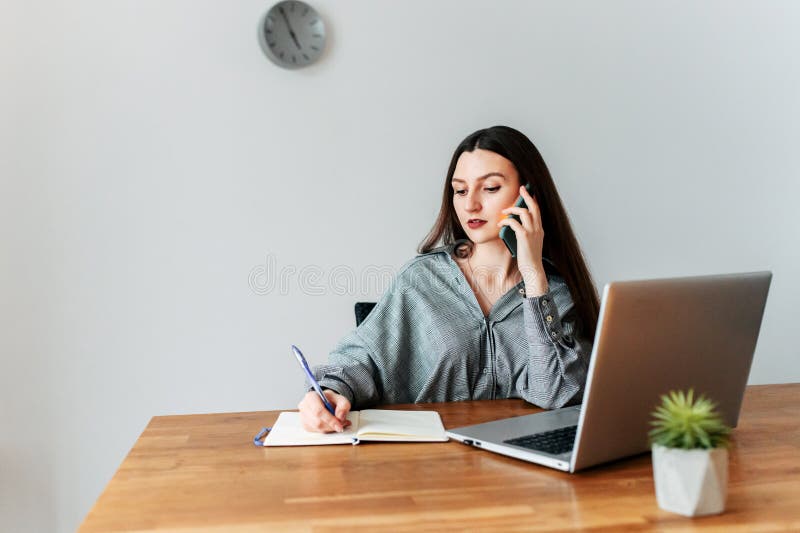 Young attractive woman in smart casual in office. She speaks on the phone and writes something in a notebook. Laptop on the table. Young attractive woman in smart casual in office. She speaks on the phone and writes something in a notebook. Laptop on the table