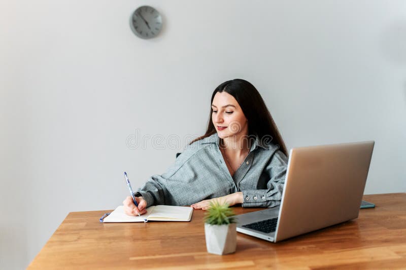 Young attractive girl student writes something in a notebook, she watches lectures on a laptop. Young attractive girl student writes something in a notebook, she watches lectures on a laptop