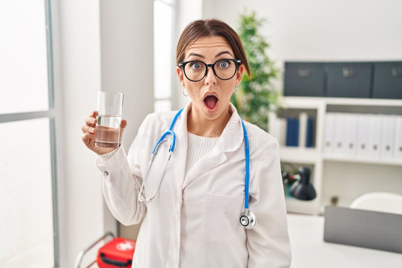 Young brunette doctor woman holding glass of water scared and amazed with open mouth for surprise, disbelief face. Young brunette doctor woman holding glass of water scared and amazed with open mouth for surprise, disbelief face