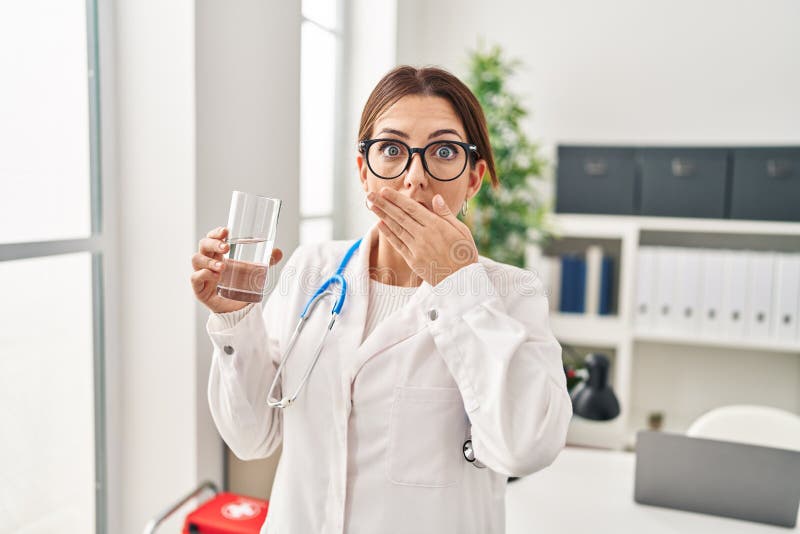 Young brunette doctor woman holding glass of water covering mouth with hand, shocked and afraid for mistake. surprised expression. Young brunette doctor woman holding glass of water covering mouth with hand, shocked and afraid for mistake. surprised expression