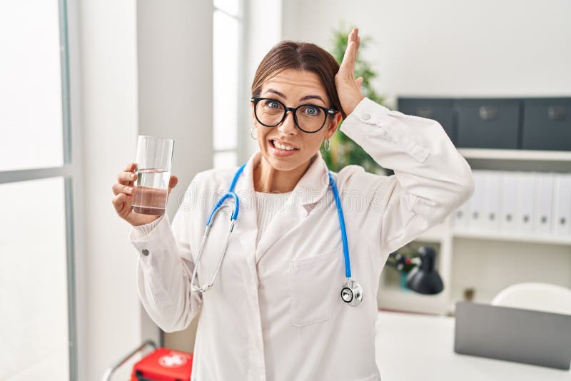 Young brunette doctor woman holding glass of water stressed and frustrated with hand on head, surprised and angry face. Young brunette doctor woman holding glass of water stressed and frustrated with hand on head, surprised and angry face