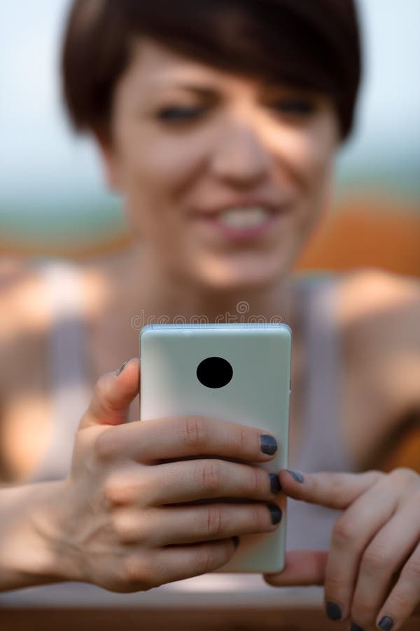 Young woman reading a message or watching media on a smartphone outdoors in dappled sunlight with focus to the phone. Young woman reading a message or watching media on a smartphone outdoors in dappled sunlight with focus to the phone