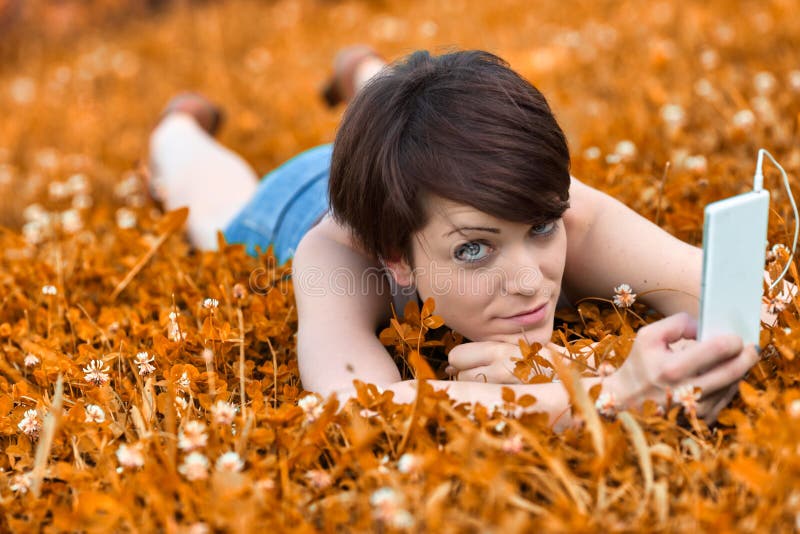 Young woman lying in a meadow listening to music on her mobile phone glancing up to keep a wary eye on the camera. Young woman lying in a meadow listening to music on her mobile phone glancing up to keep a wary eye on the camera