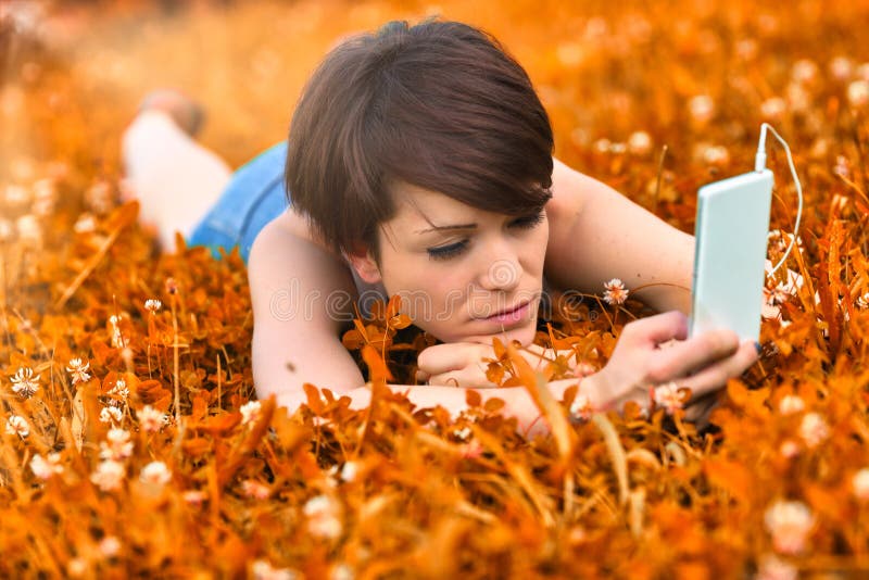 Young woman lying in a meadow selecting a tune on her mobile phone as she spends a relaxing day listening to music. Young woman lying in a meadow selecting a tune on her mobile phone as she spends a relaxing day listening to music