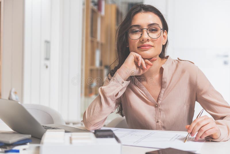 Talented female designer is planning new project. She is sitting at desk near blueprint and smiling. Talented female designer is planning new project. She is sitting at desk near blueprint and smiling