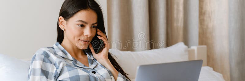 Młoda kobieta rozmawiająca przez telefon komórkowy i używająca laptopa leżąc w łóżku