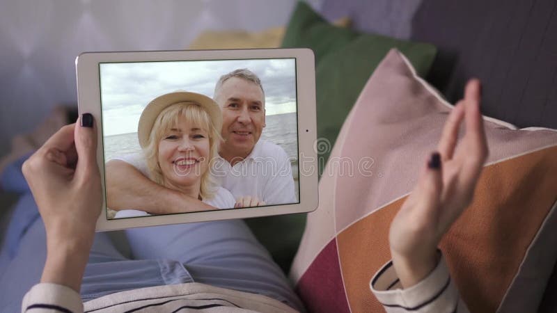 Młoda kobieta rozmawiająca ze swoimi szczęśliwymi rodzicami przez wideorozmowę z tabletem, leżąc na sofie