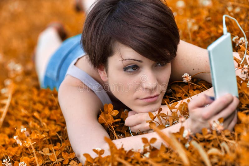 Young woman listening to music or watching media on a mobile phone as she relaxes in a meadow amongst the wildflowers. Young woman listening to music or watching media on a mobile phone as she relaxes in a meadow amongst the wildflowers