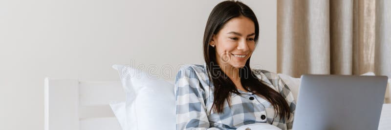 Młoda latynoska uśmiechnięta i używająca laptopa leżąc w łóżku