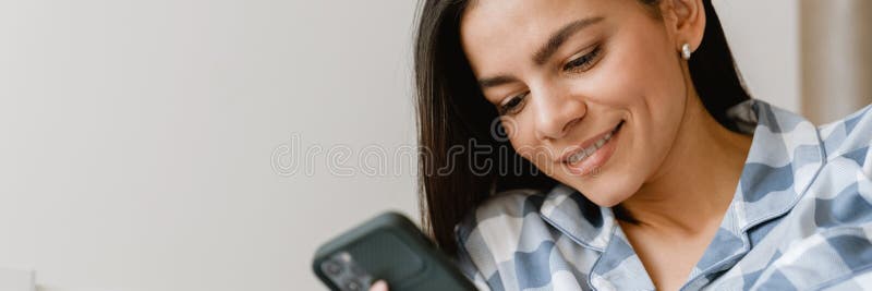Młoda latynoska uśmiechnięta i używająca telefonu komórkowego leżąc w łóżku