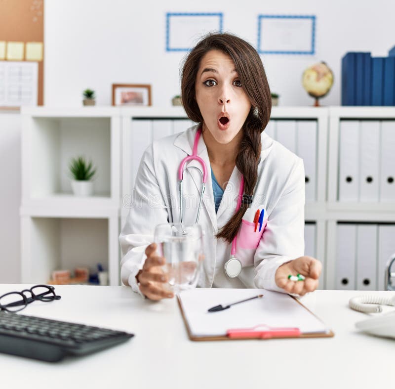 Young doctor woman holding glass of water and prescription pills afraid and shocked with surprise and amazed expression, fear and excited face. Young doctor woman holding glass of water and prescription pills afraid and shocked with surprise and amazed expression, fear and excited face