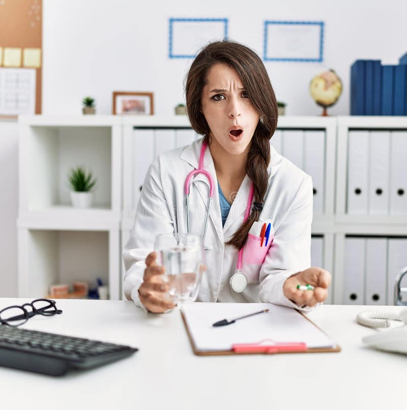 Young doctor woman holding glass of water and prescription pills in shock face, looking skeptical and sarcastic, surprised with open mouth. Young doctor woman holding glass of water and prescription pills in shock face, looking skeptical and sarcastic, surprised with open mouth