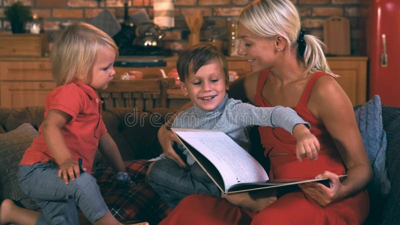 Młoda matka siedzi na kanapie z dwiema dziećmi, chłopcami, książką w rękach, starszy brat coś mówi w jej uchu