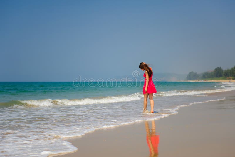 Młoda nikła długowłosa brunetka w czerwieni sukni chodzi bosego na tropikalnej plaży wzdłuż kipieli przeciw niebieskiemu niebu i fotografia stock Młoda nikła długowłosa brunetka w czerwieni sukni chodzi bosego na tropikalnej plaży wzdłuż kipieli przeciw niebieskiemu niebu i fotografia stock