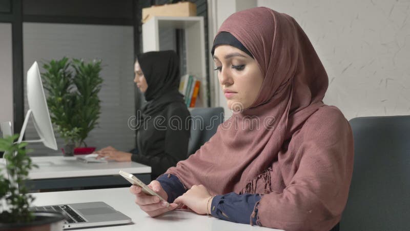 Młoda piękna dziewczyna w różowym hijab siedzi w biurze i używa smartphone Dziewczyna w czarnym hijab w tle arabskie kobiety