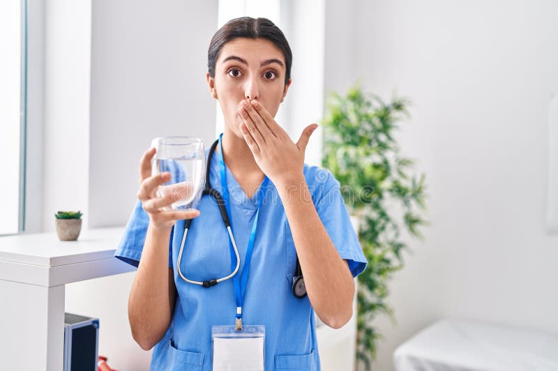 Young beautiful woman wearing doctor uniform and stethoscope drinking water covering mouth with hand, shocked and afraid for mistake. surprised expression. Young beautiful woman wearing doctor uniform and stethoscope drinking water covering mouth with hand, shocked and afraid for mistake. surprised expression