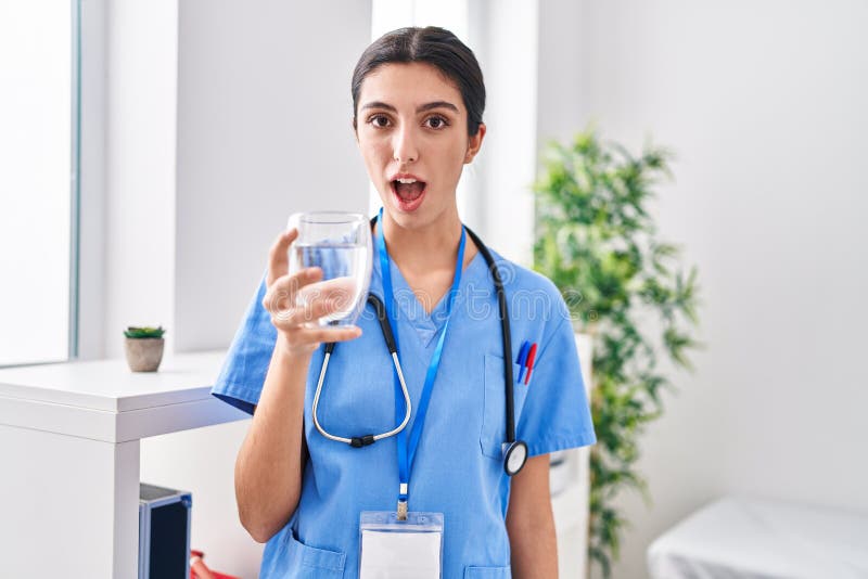 Young beautiful woman wearing doctor uniform and stethoscope drinking water scared and amazed with open mouth for surprise, disbelief face. Young beautiful woman wearing doctor uniform and stethoscope drinking water scared and amazed with open mouth for surprise, disbelief face