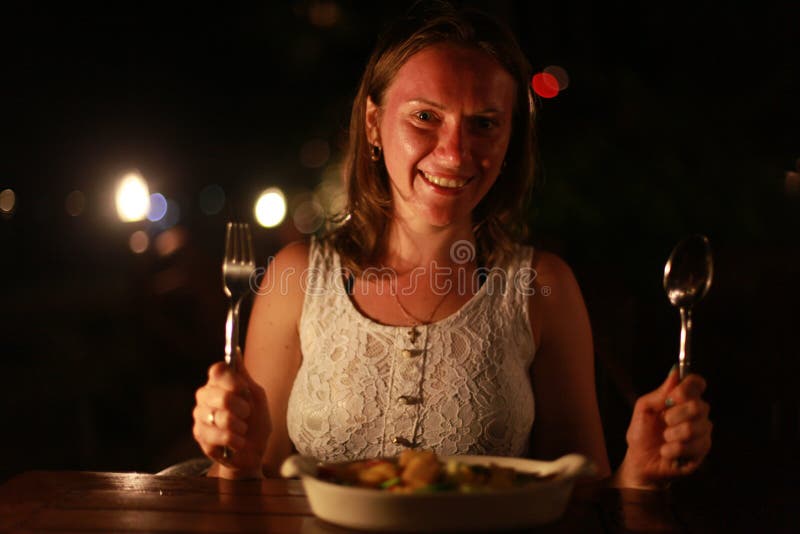 Młoda rozochocona uśmiechnięta kobiety mienia łyżka i rozwidlenie w plenerowej kawiarni przy morzem na pięknych bokeh światłach fotografia stock Młoda rozochocona uśmiechnięta kobiety mienia łyżka i rozwidlenie w plenerowej kawiarni przy morzem na pięknych bokeh światłach fotografia stock