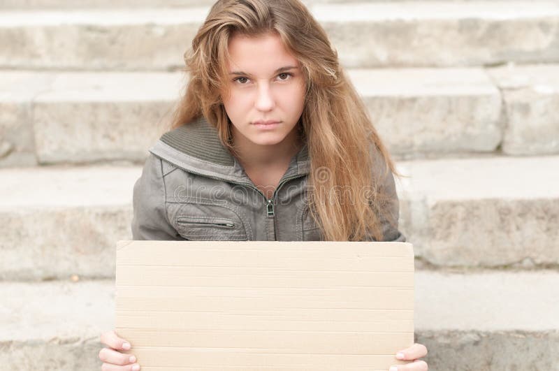 Abandoned teenage girl sitting outdoor on grey stone steps with blank cardboard sign in her hands. Young girl looking sad and depressed. Neglected teens. Beggar and tramp. Unemployment. Abandoned teenage girl sitting outdoor on grey stone steps with blank cardboard sign in her hands. Young girl looking sad and depressed. Neglected teens. Beggar and tramp. Unemployment.