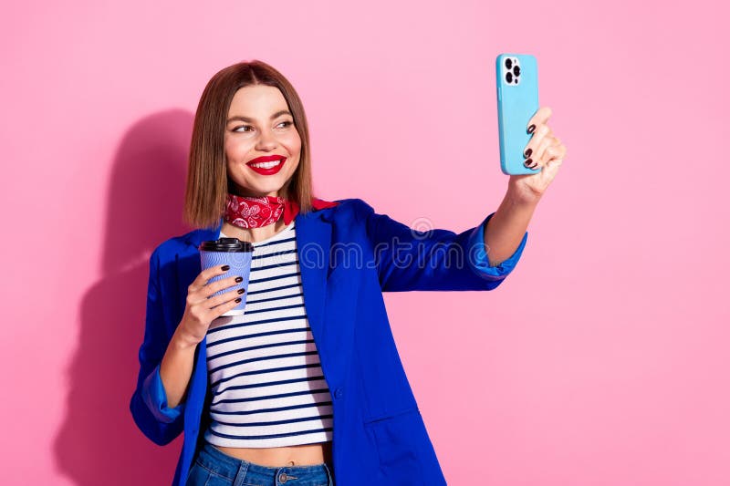 Młoda stylowa kobieta w niebieskiej kurtce zabierająca selfie trzymając filiżankę kawy na różowym tle pokazującym fotografia royalty free