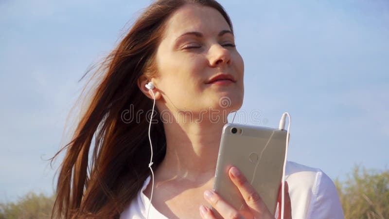 Młoda szczęśliwa kobieta używa wiszącą ozdobę przeciw niebieskiemu niebu Żeńska słuchająca muzyka przez telefonu komórkowego w zw