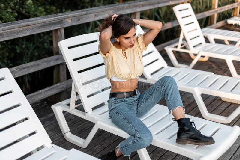 Młoda śliczna kobieta modelka w modnych jeansach z butami i żółtą bluzką mody siedzi na łóżku słonecznym na drewnianym molo zdjęcia royalty free