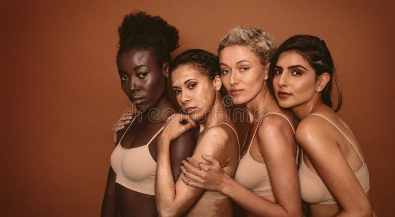 Portrait of young women with different skin types on brown background. Diverse group of females standing together and looking at camera. Portrait of young women with different skin types on brown background. Diverse group of females standing together and looking at camera.