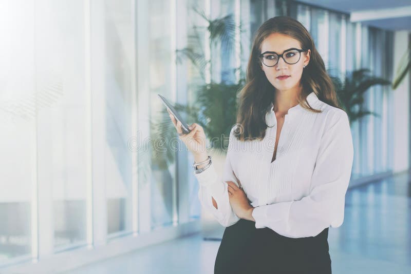 Młody atrakcyjny bizneswoman w szkłach i białej koszula stoi w biurowym, używać smartphone zdjęcia stock Młody atrakcyjny bizneswoman w szkłach i białej koszula stoi w biurowym, używać smartphone zdjęcia stock