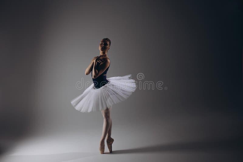 Young ballerina in elegance white tutu and pointe shoes dancing against dark background. Graceful litted ballerina performing in dark. Ballet art, grace, flexibility, motion. Young ballerina in elegance white tutu and pointe shoes dancing against dark background. Graceful litted ballerina performing in dark. Ballet art, grace, flexibility, motion