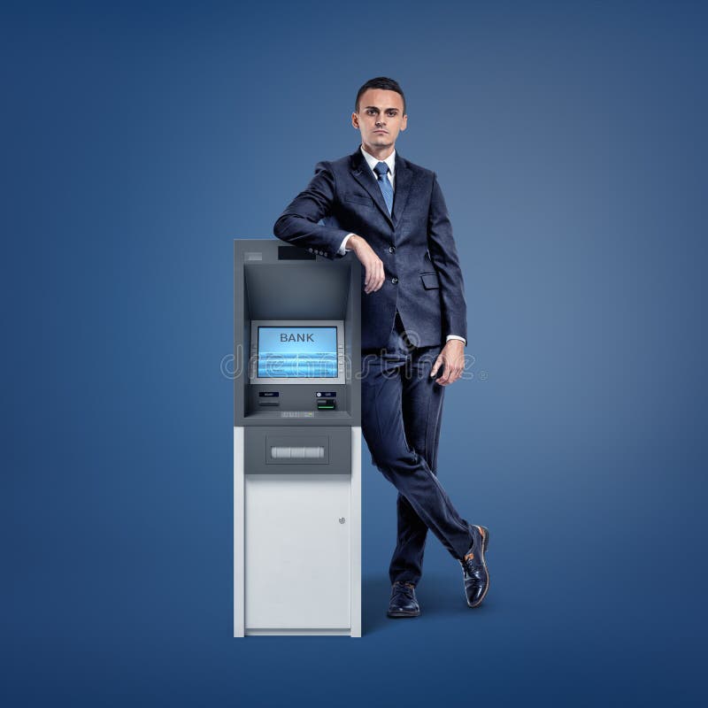 Młody biznesmen opiera na małej banka ATM maszynie na zmroku - błękitny tło obrazy royalty free Młody biznesmen opiera na małej banka ATM maszynie na zmroku - błękitny tło obrazy royalty free