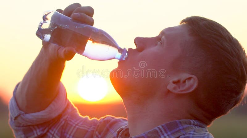 Młody człowiek pije jasno wodę od plastikowej butelki w naturze przy pszenicznym polem Mężczyzna ma wodną przerwę przy zmierzchem