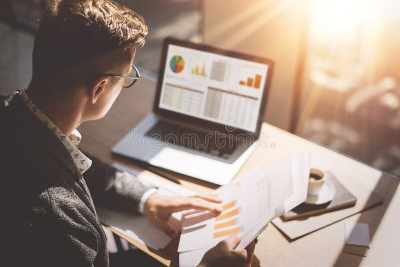 Młody finansowy targowy analityk w eyeglasses pracuje przy pogodnym biurem na laptopie podczas gdy siedzący przy drewnianym stołe Młody finansowy targowy analityk w eyeglasses pracuje przy pogodnym biurem na laptopie podczas gdy siedzący przy drewnianym stołe