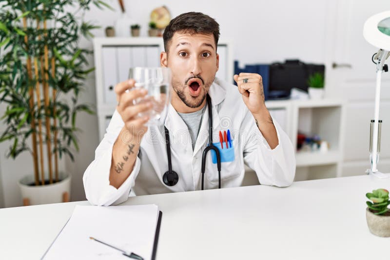 Young doctor holding glass of water scared and amazed with open mouth for surprise, disbelief face. Young doctor holding glass of water scared and amazed with open mouth for surprise, disbelief face