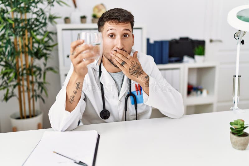 Young doctor holding glass of water covering mouth with hand, shocked and afraid for mistake. surprised expression. Young doctor holding glass of water covering mouth with hand, shocked and afraid for mistake. surprised expression