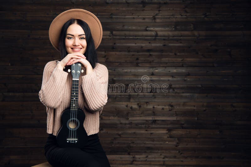 Młody piękny kobiety obsiadanie na krześle jest ubranym kapelusz z ukulele Dziewczyna z rękami składał na małej gitarze na zmroku obraz royalty free Młody piękny kobiety obsiadanie na krześle jest ubranym kapelusz z ukulele Dziewczyna z rękami składał na małej gitarze na zmroku obraz royalty free