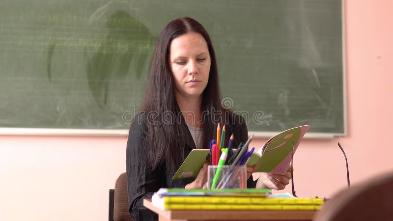 Młody, piękny nauczyciel siedzi przed tablicą i patrzy w notes.